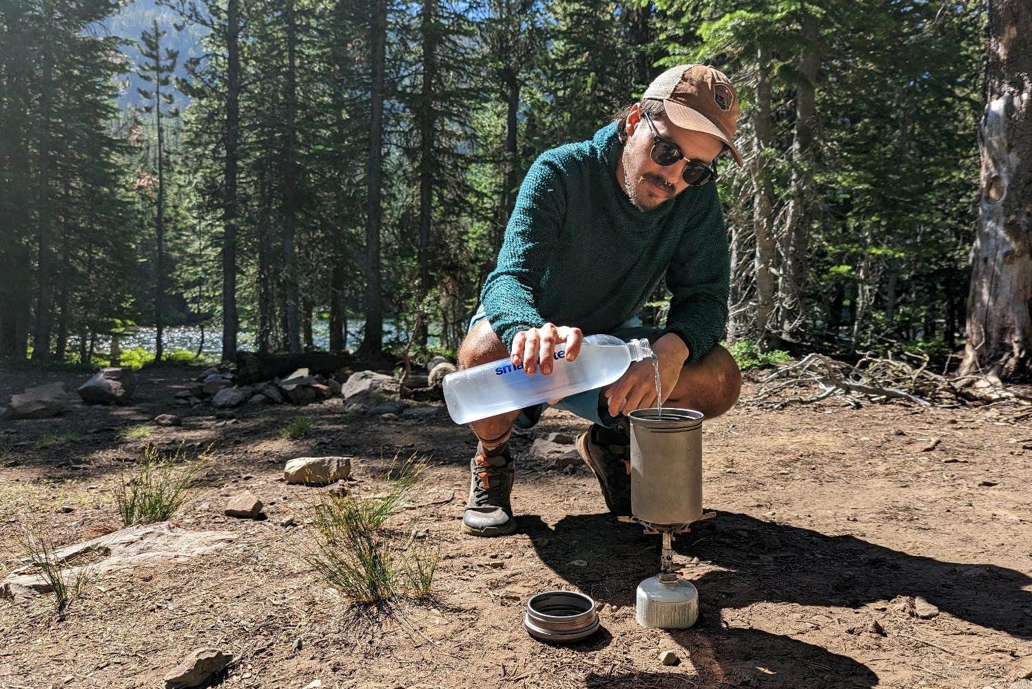 A hiker pouring water into a cookpot on top of the SOTO Windmaster stove - hes in a campsite surrounded by pine trees with a lake in the background