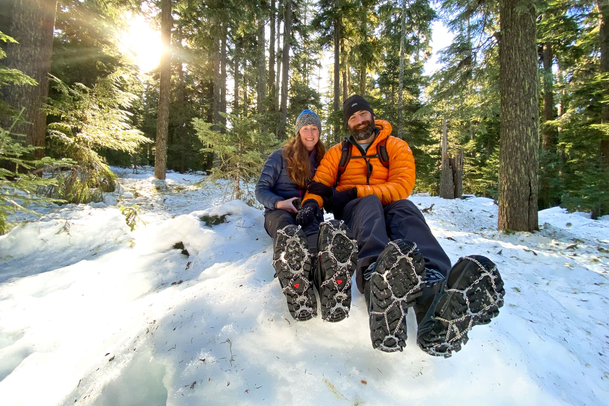 A couple wearing the Kahtoola MICROspikes and Snowline Chainsen Trail Light traction devices on a snowy winter hike in the woods