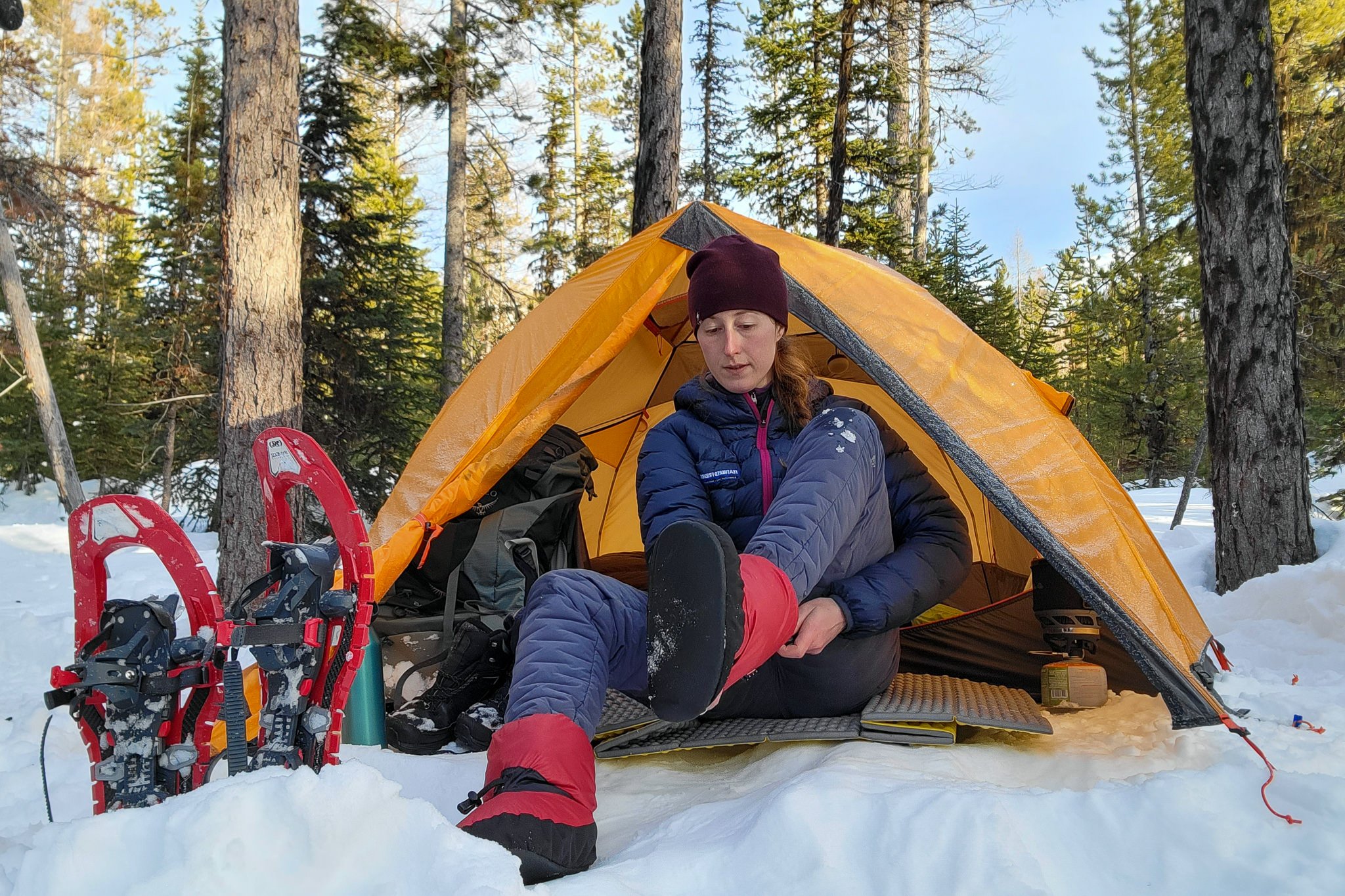 A hiker putting on down booties in the doorway of a tent with TSL Symbioz Hyperflex elite Snowshoes next to them