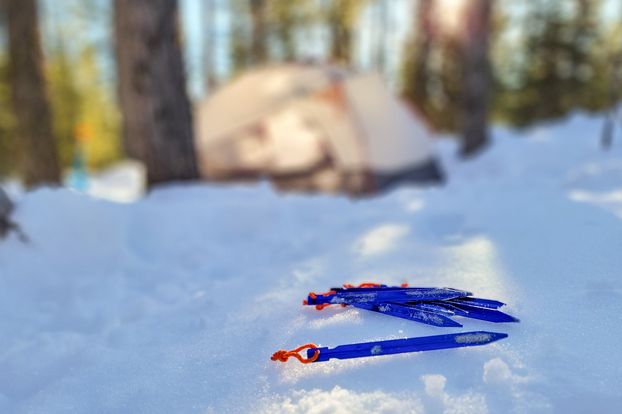 A set of All One tech tent stakes laying in the snow with a tent in the background