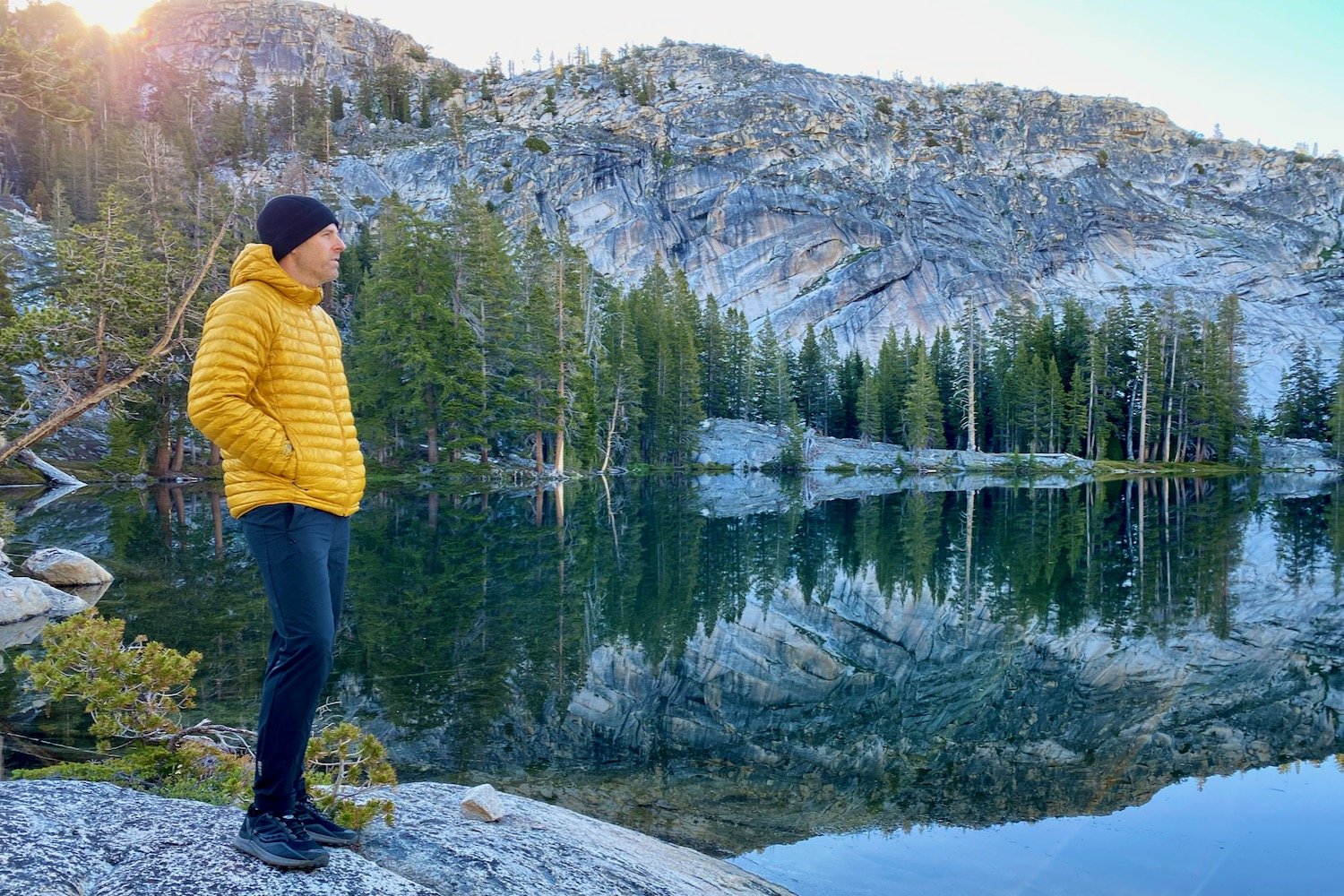 A backpacker standing near granite-lined lake in a yellow Mountain Hardwear Ghost Whisperer UL down jacket