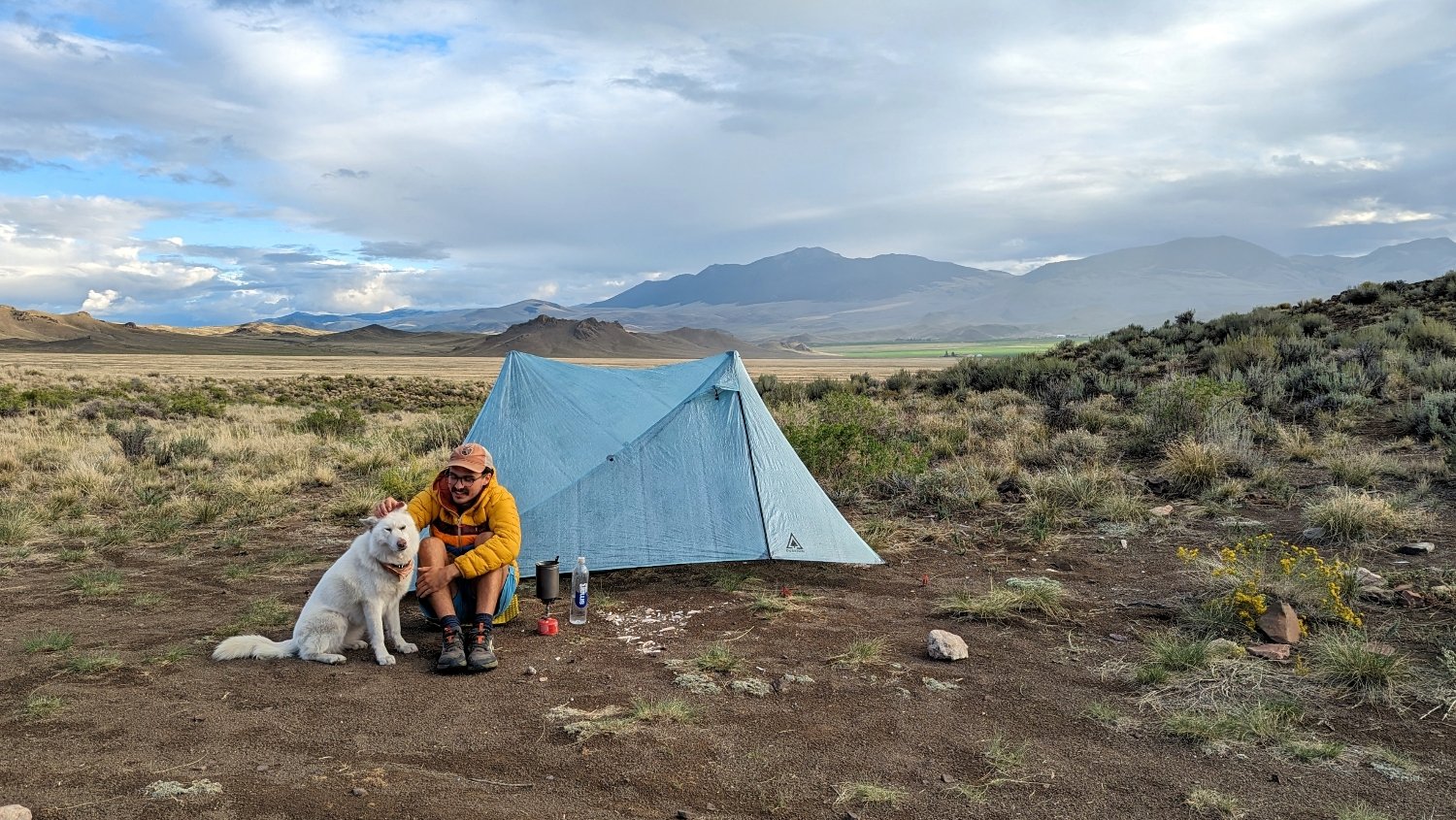 A hiker and a dog sitting outside in a campsite in front of the Durston X-Mid 2 Pro tent. There are mountains in the background with rainclouds overhead