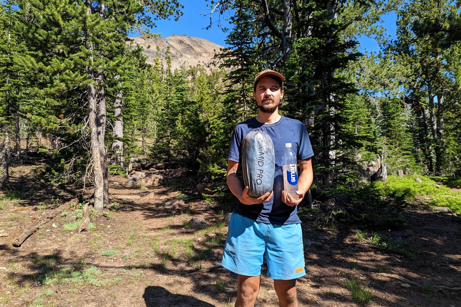 A hiker holding the Durston X-Mid 2 Pro tent packed in its stuff sack in one hand a Smartwater bottle in the other hand to show the size comparison of the two. There's a mountain peak peaking out from behind some pine trees in the background