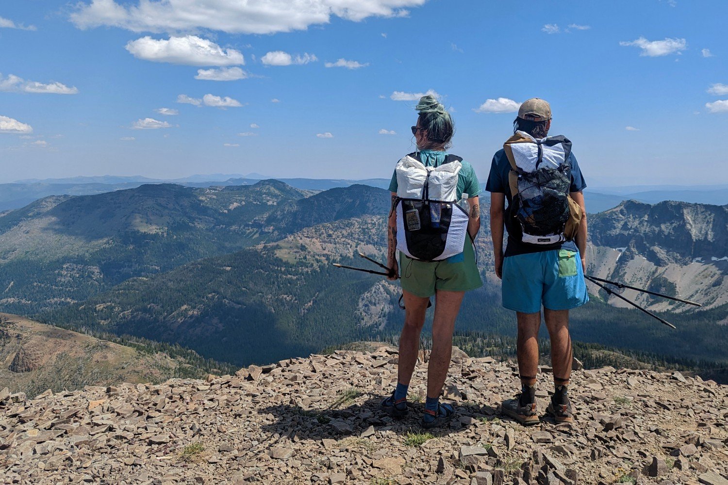 Two hikers with ultralight backpacks standing at the peak of a mountain looking out at more distant mountain peaks