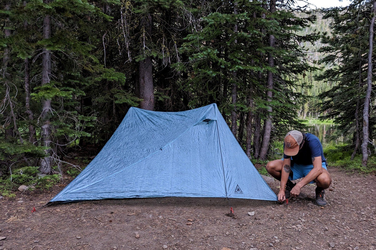 A hiker staking down the Durston X-Mid 2 Pro tent in a campsite surrounded by pine trees