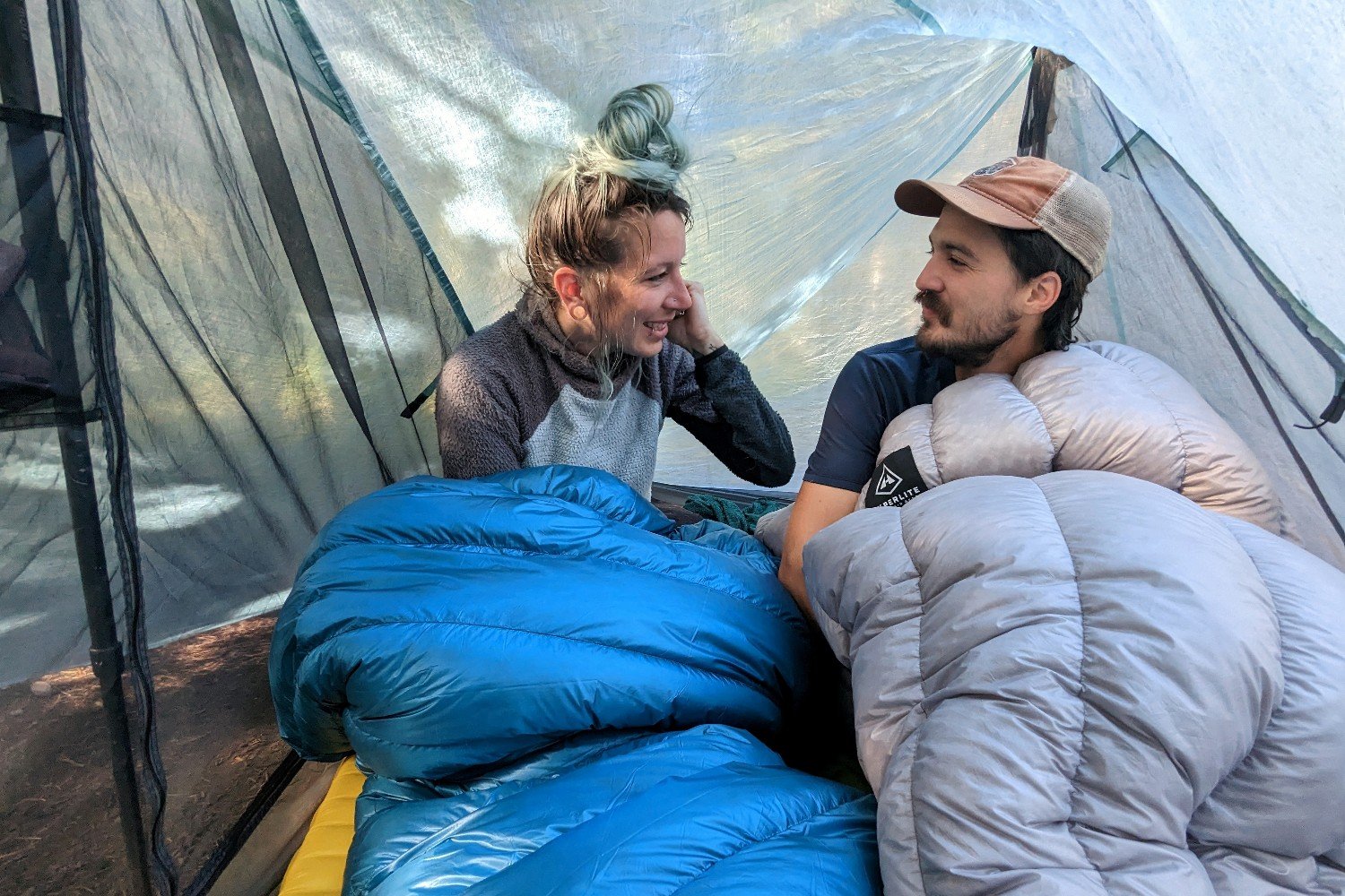 An interior view of the Durston X-Mid 2 Pro tent. Two hikers are sitting up wrapped up in quilts. The hiker onthe right has plenty of headroom and the hiker on the left is leaning down away from the wall because there's less headroom