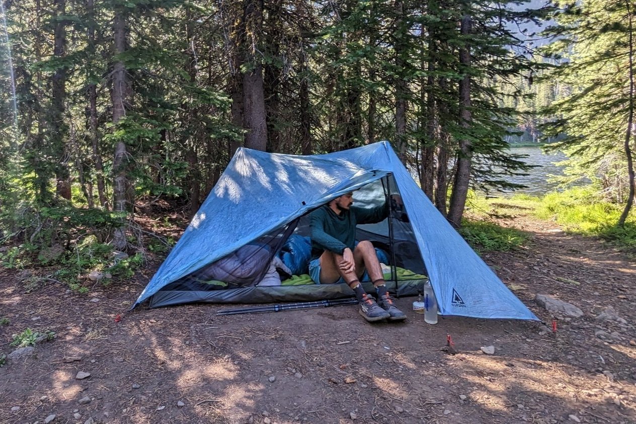A hiker sitting inside the Durston X-Mid 2 Pro tent with front door and vestibule open. There are pine trees and a glimpse of a lake in the background