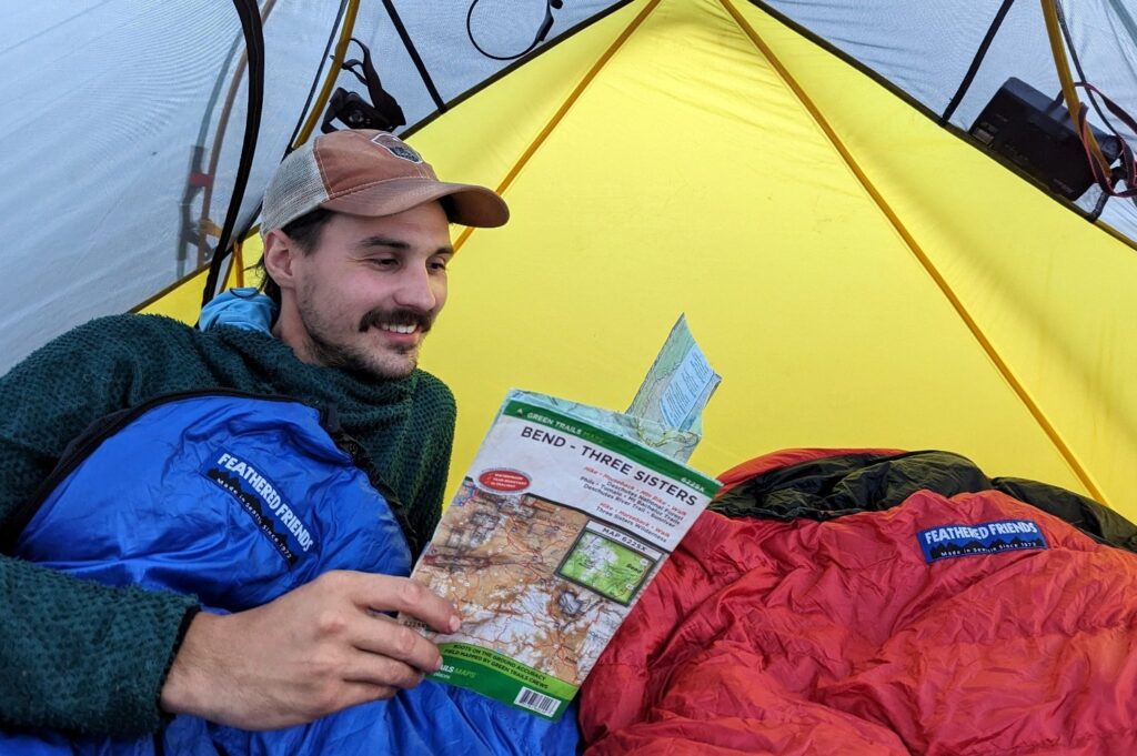 A man reads inside a blue sleeping bag inside a tent while backpacking.