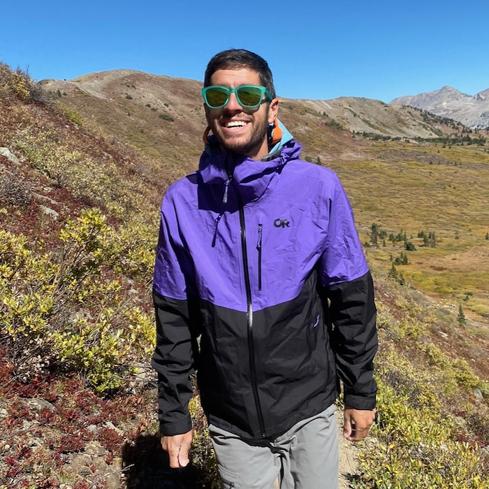 Against a summer mountain backdrop in the Colorado Rockies, a man in purple and black rain jacket and green sunglasses hikes towards camera while smiling