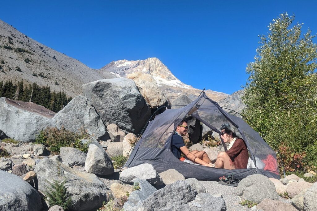 Two hikers sitting inside the Sea to Summit Telos TR2 in a rocky campsite with the doors open. The hikers are looking at each other and smiling, and there's a mountain peak in the background