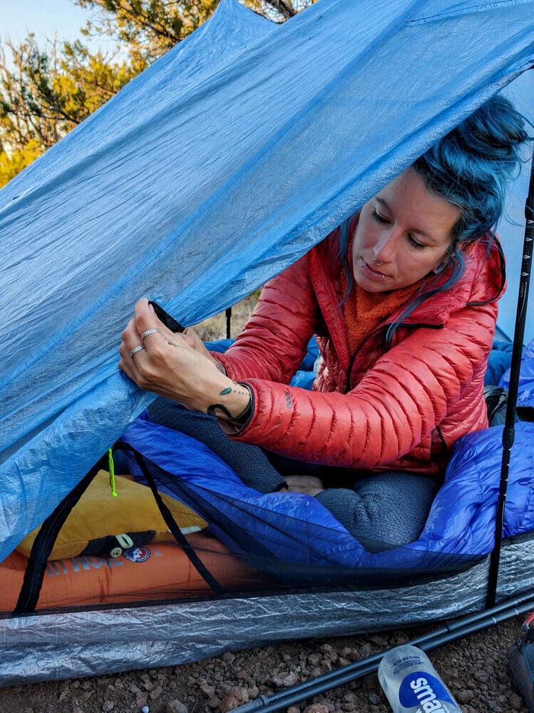 A hiker adjusting the doors of the Zpacks Duplex Zip tent from the inside while in a sleeping bag sitting on a sleeping pad in the Arizona wilderness