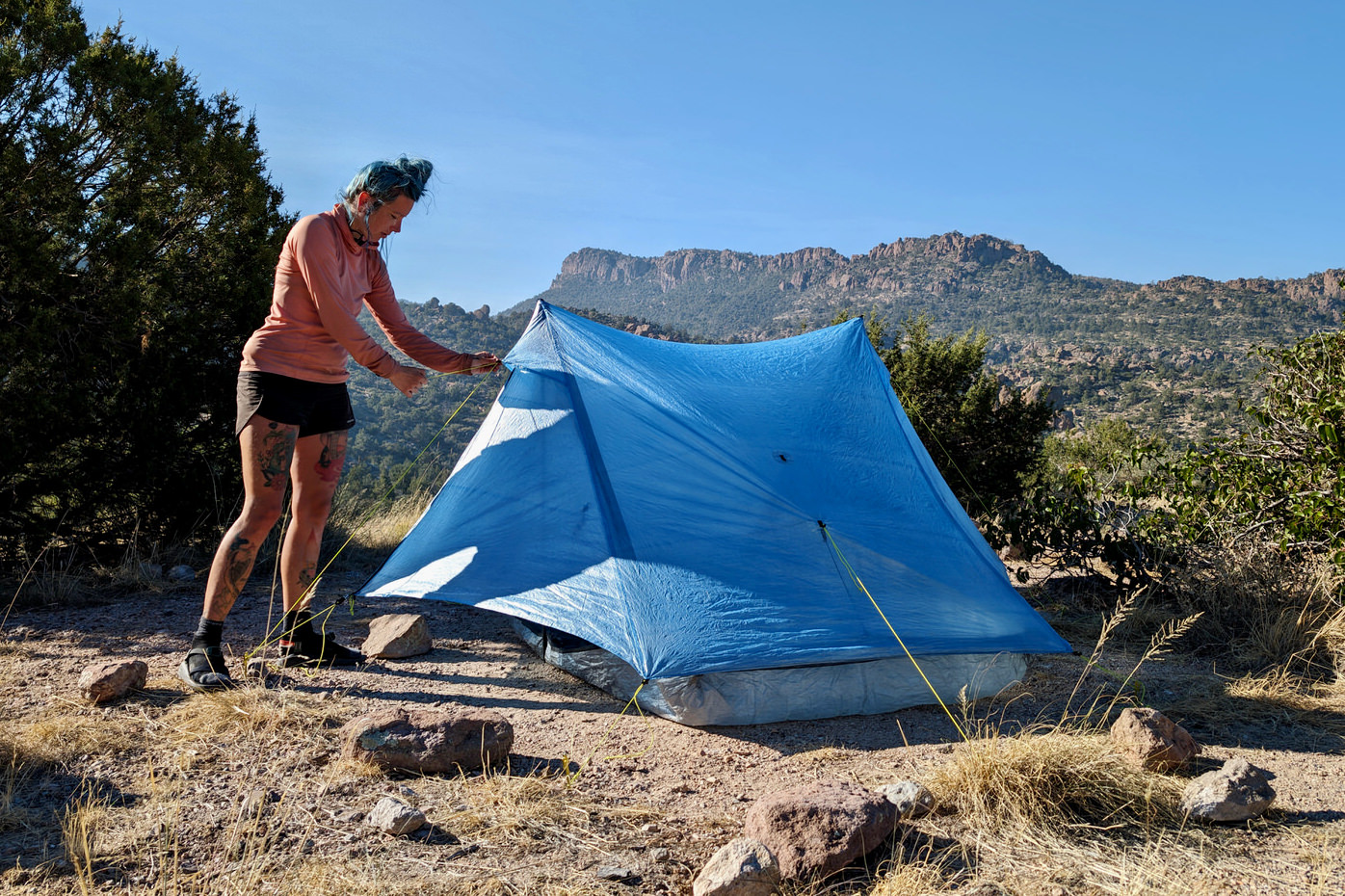 A backpacker adjusting the left outside guyline of the Zpacks Duplex Zip tent in Arizona with mountains in the background and blue sky overhead