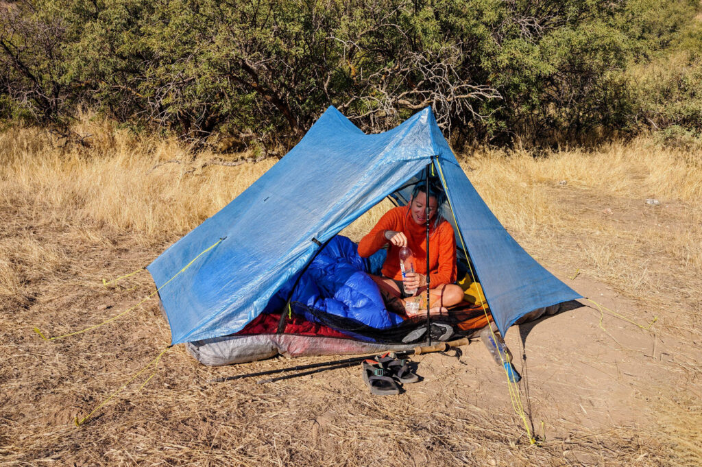 A backpacker sitting inside the Zpacks Duplex Zip tent in a sunny field with desert trees in the background