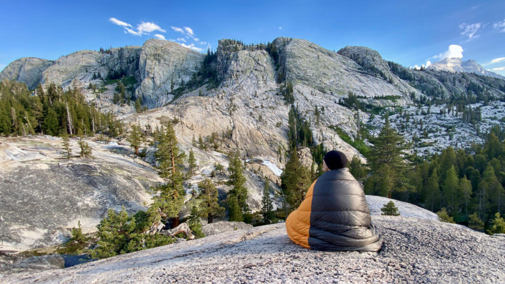 A backpacker wrapped up in the Enlightened Equipment Revelation sleeping quilt while sitting on a rocky slab overlooking bare granite mountains across the valley