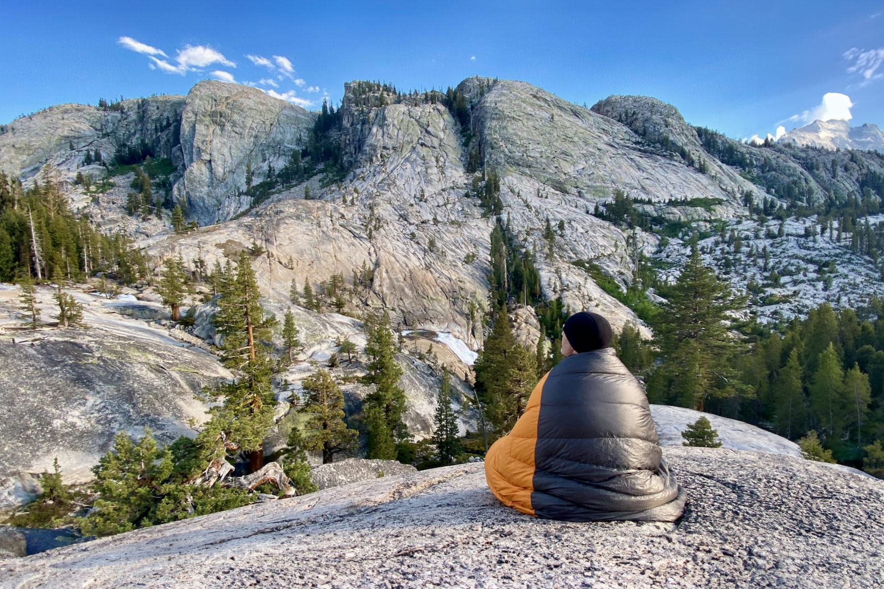 A backpacker wrapped up in the Enlightened Equipment Revelation sleeping quilt while sitting on a rocky slab overlooking bare granite mountains across the valley
