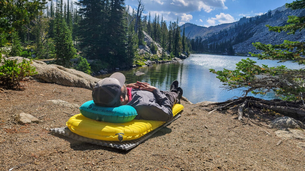 A backpacker relaxing on the shore of an alpine lake surrounded by mountains on an inflatable sleeping pad and the Sea to Summit Aeros Ultralight Pillow