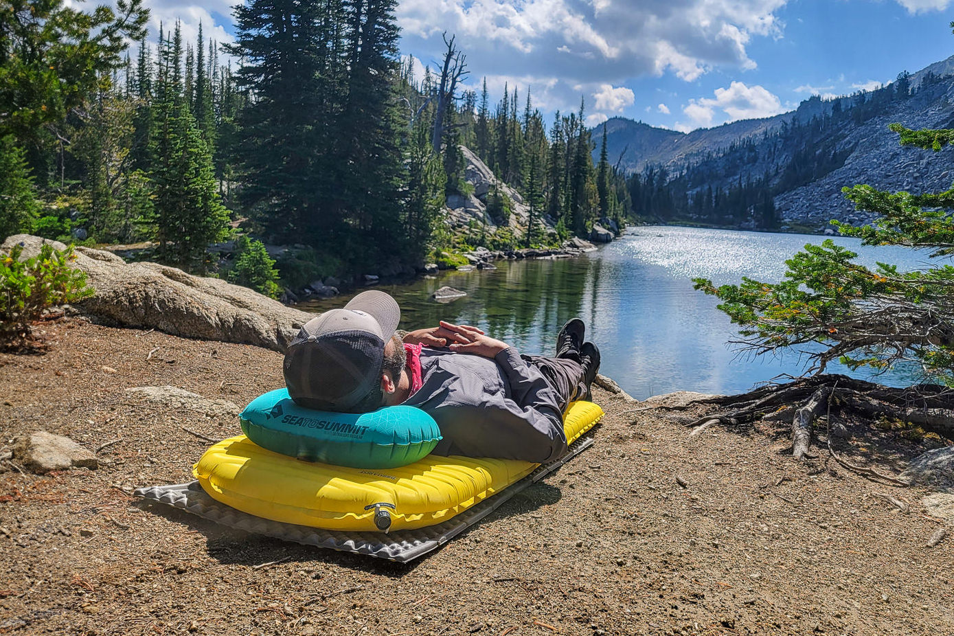 A backpacker relaxing on the shore of an alpine lake surrounded by mountains on an inflatable sleeping pad and the Sea to Summit Aeros Ultralight Pillow