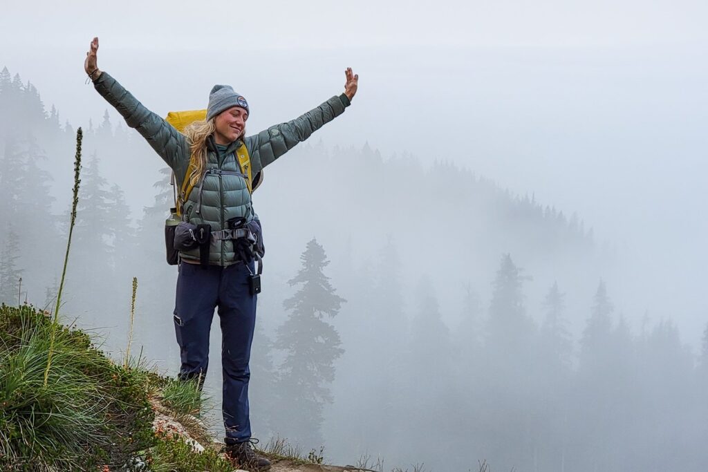 A smiling hiker standing on a foggy trail with obscured pine trees in the background. She has her arms stretched in the air and she's wearing an REI 650 Down Jacket
