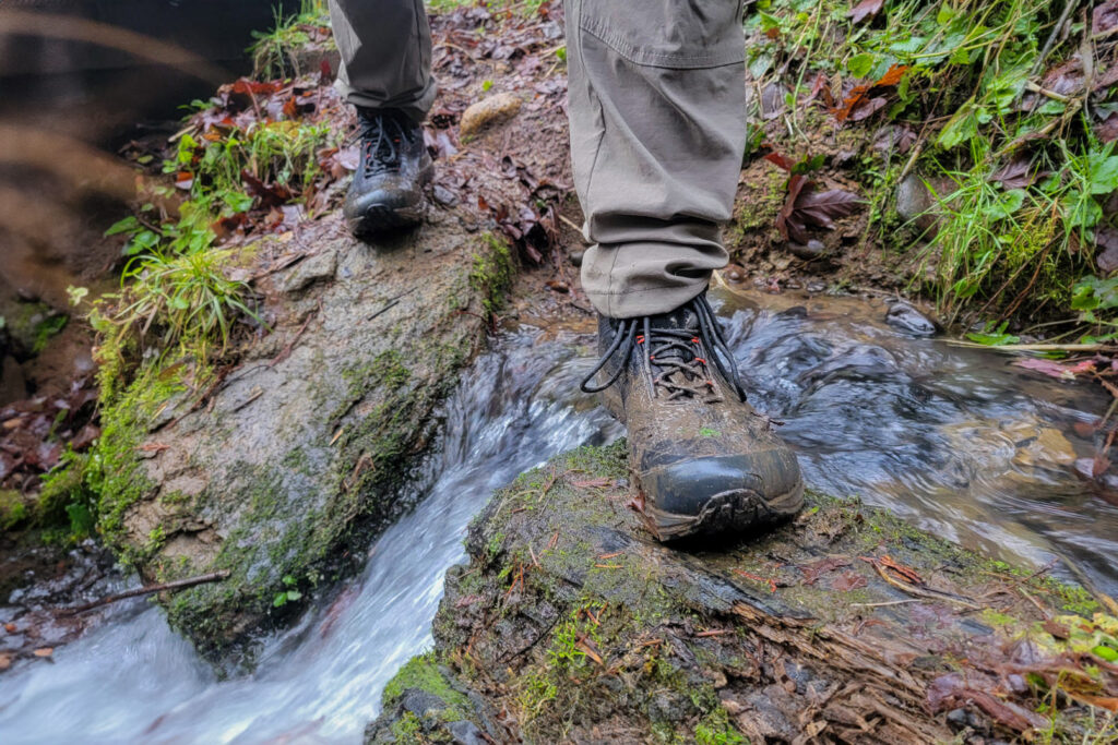 Closeup of a hiker's feet crossing a creek in a muddy pair of Topo Athletic Trailventure 2 hiking boots