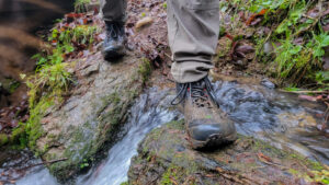 Closeup of a hiker's feet crossing a creek in a muddy pair of Topo Athletic Trailventure 2 hiking boots