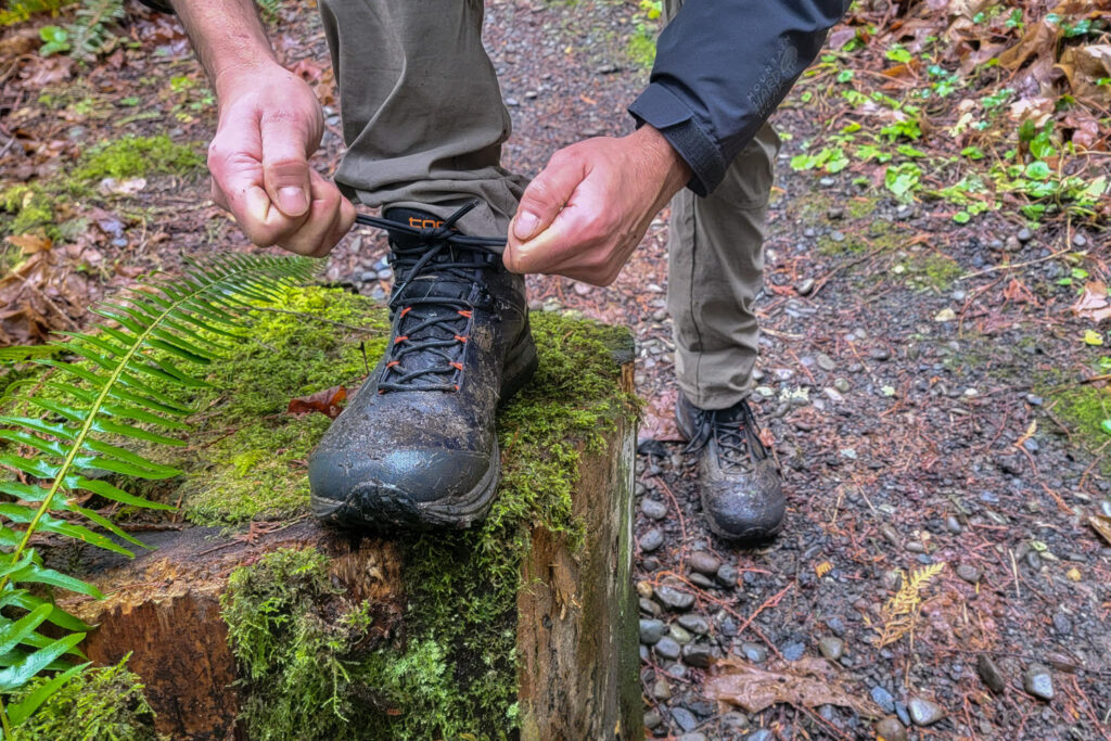 A hiker tying the laces of his muddy Topo Athletic Trailventure 2 boot up on a mossy stump