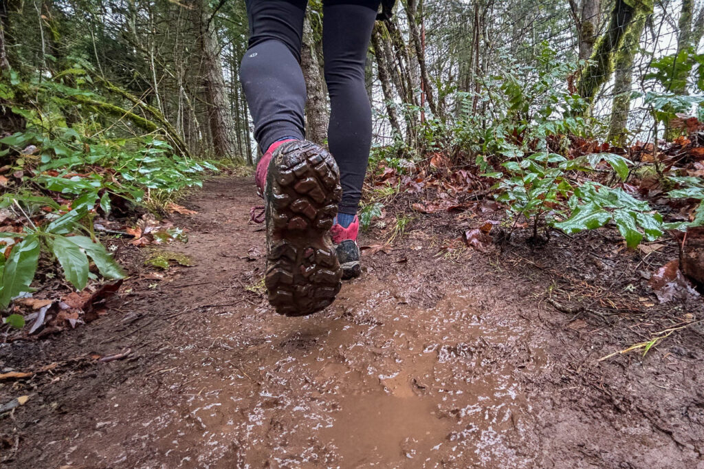 View of the sole of hiker's dirty Obōz Bridger Waterproof hiking boot and they walk down a muddy trail in the woods
