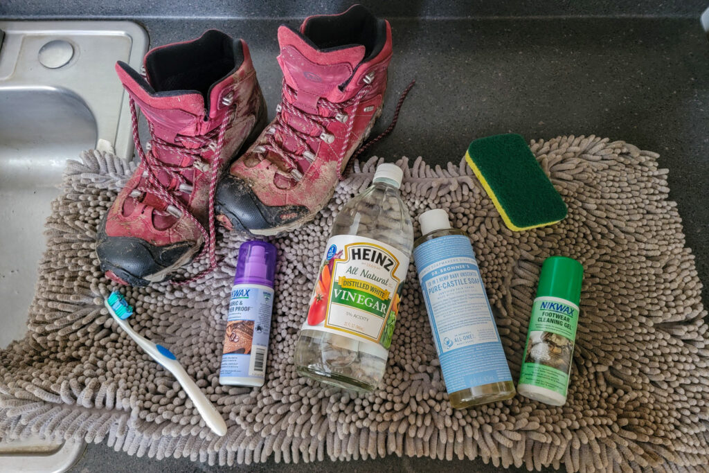 A pair of muddy Obōz Bridger hiking boots on the kitchen counter with cleaning supplies including a toothbrush, Nikwax Fabric & Leather Proof, white vinegar, Nikwax Footwear Cleaning Gel, Dr. Bronner's Castile Soap, and a sponge