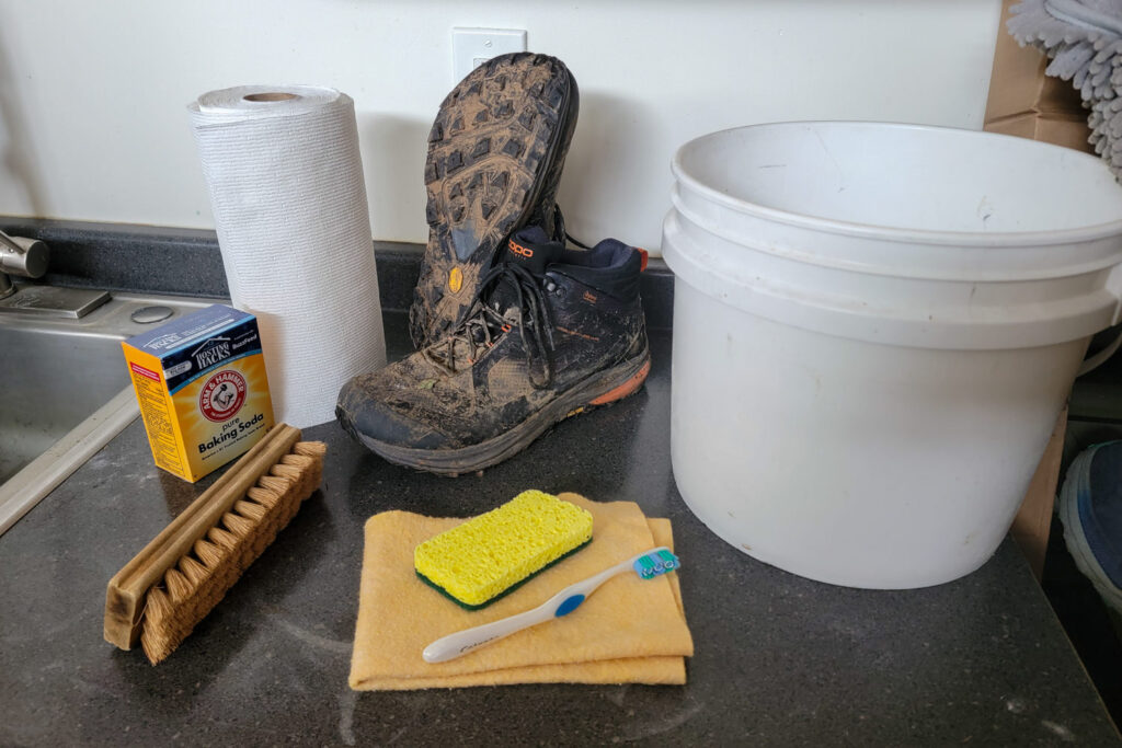 A pair of dirty Topo Athletic Trailventure 2 hiking boots on the kitchen counter with a bucket, rag, sponge, toothbrush, bristle brush, baking soda, and paper towels
