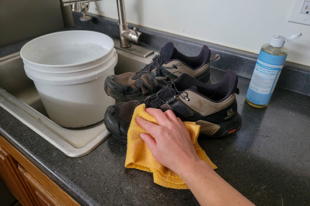 A person spot cleaning a pair of the Salomon X Ultra hiking shoes with a rag and diluted Dr. Bronner's Castile Soap