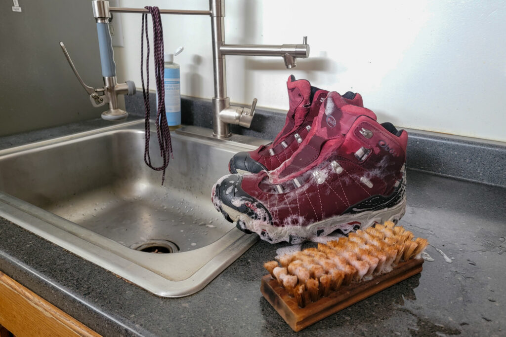 A brush and pair of sudsy red Obōz Bridger WP hiking boots next to a kitchen sink with the laces hanging in the background