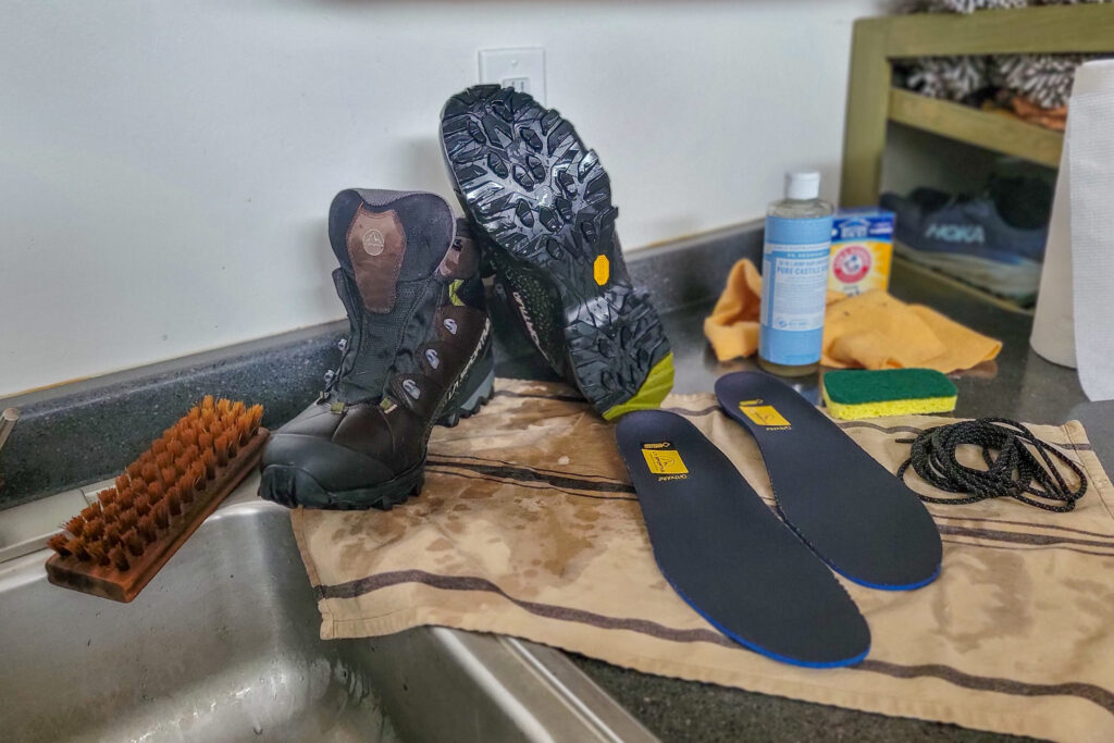 A pair of La Sportiva Nucleo High II GTX hiking boots next to a sink for cleaning with a brush, soap, baking soda, a rag, sponge, and towel