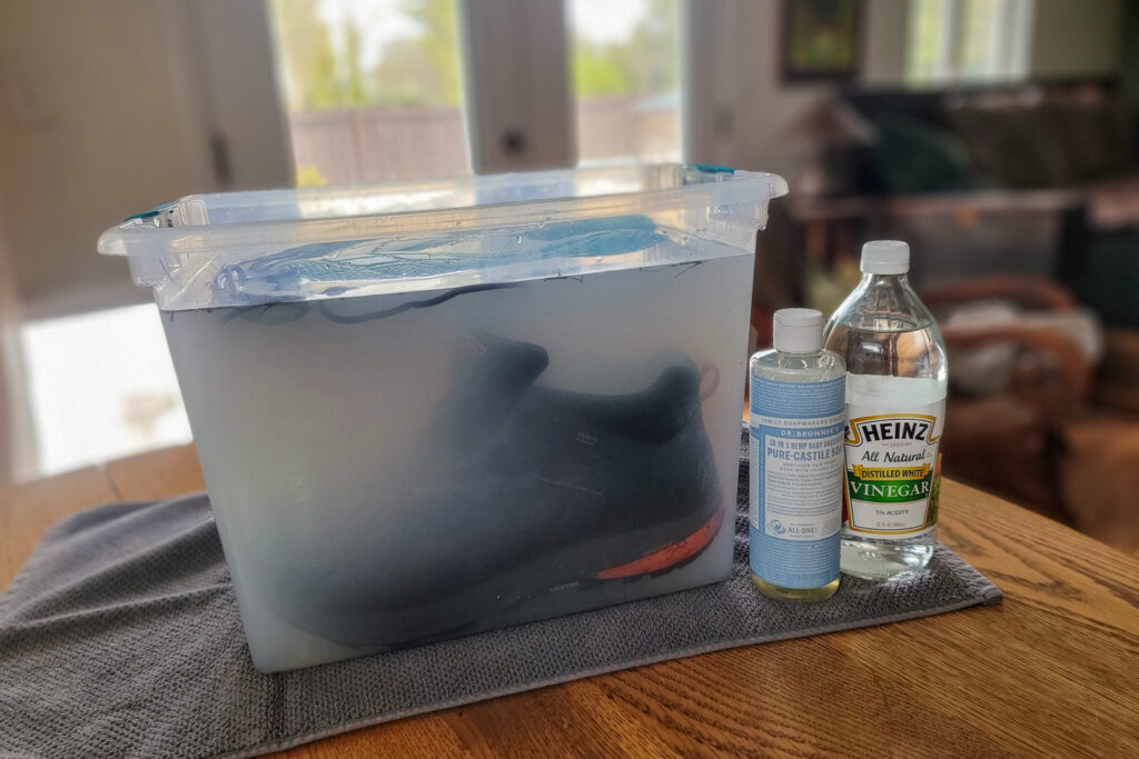 A pair of Topo Athletic Trailventure 2 soaking in a clear storage box full of water with bottles of Dr. Bronner's Castile Soap and white vinegar beside it on the table