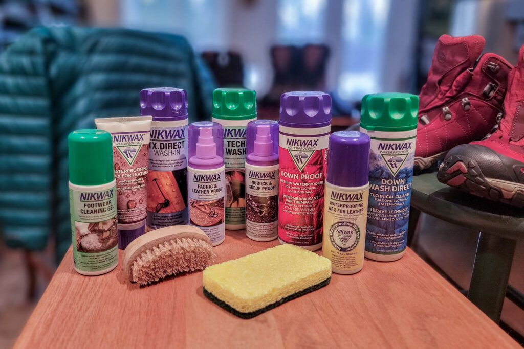 Nikwax Products lined up on a wooden table with a brush, sponge, hiking boots, and a down jacket