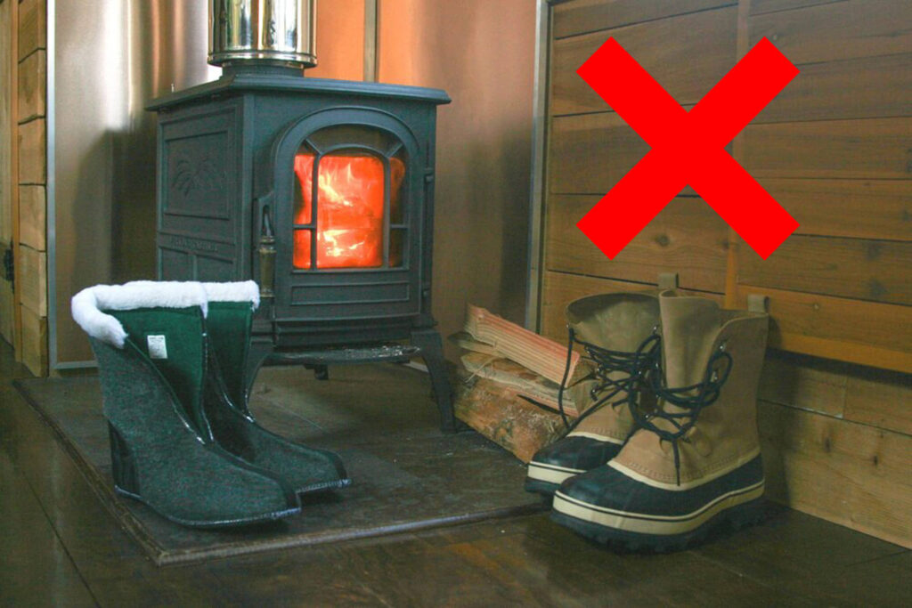 A pair of Sorel Caribou winter boots sitting next to a glowing woodstove with the liners removed with a red X in the corner
