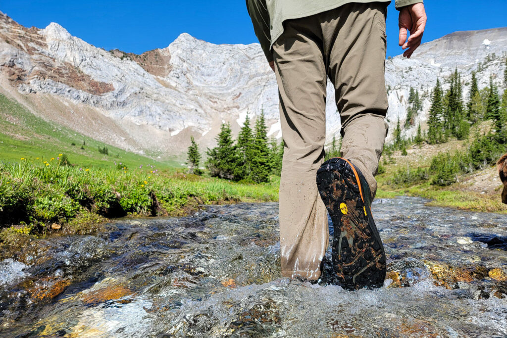 A hiker crossing a creek in the Topo Athletic Ultraventure 3 hiking shoes with a marbled mountain in the background