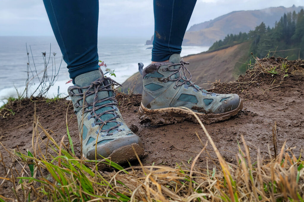 A hiker standing on a muddy trail in a pair of Vasque Breeze hiking boots with the ocean and coastal bluffs in the background