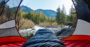 Looking out the doorway of a backpacking tent over the foot of a sleeping bag with a river and mountains in the background