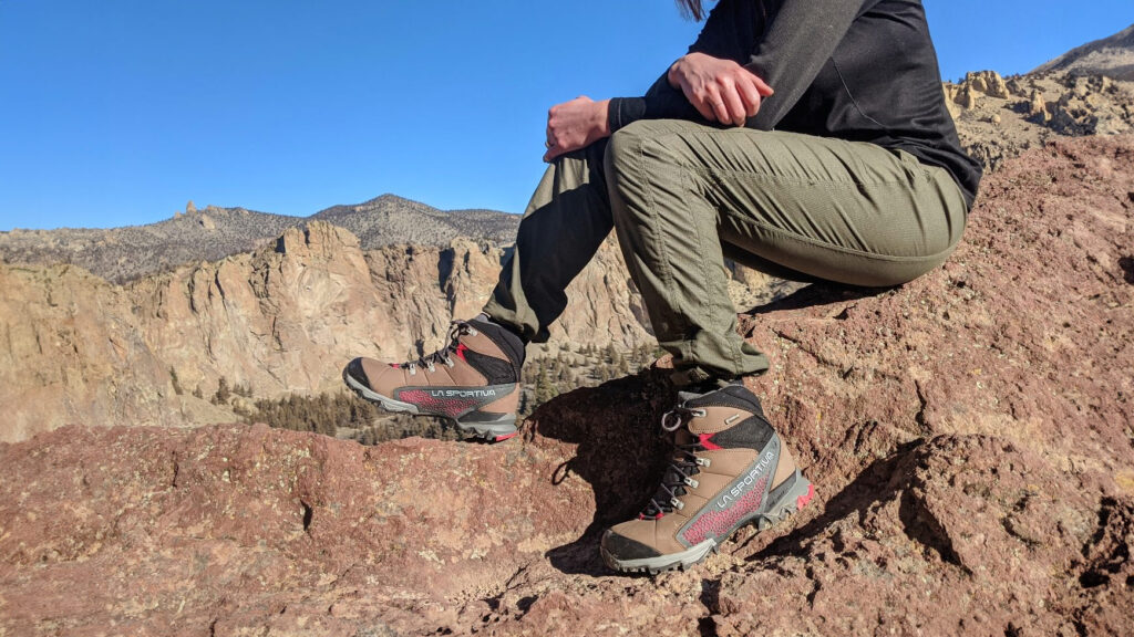 Waist-down view of a hiker taking a rest break by sitting on some rocks in the La Sportiva Nucleo High II GTX boots with rocky, high-desert clifs in the background.