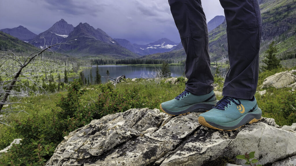 Knee-down view of a hiker standing on a white granite rock in a pair of teal Topo Athletic Ultraventure 3 hiking shoes in front a lake in Glacier National Park.