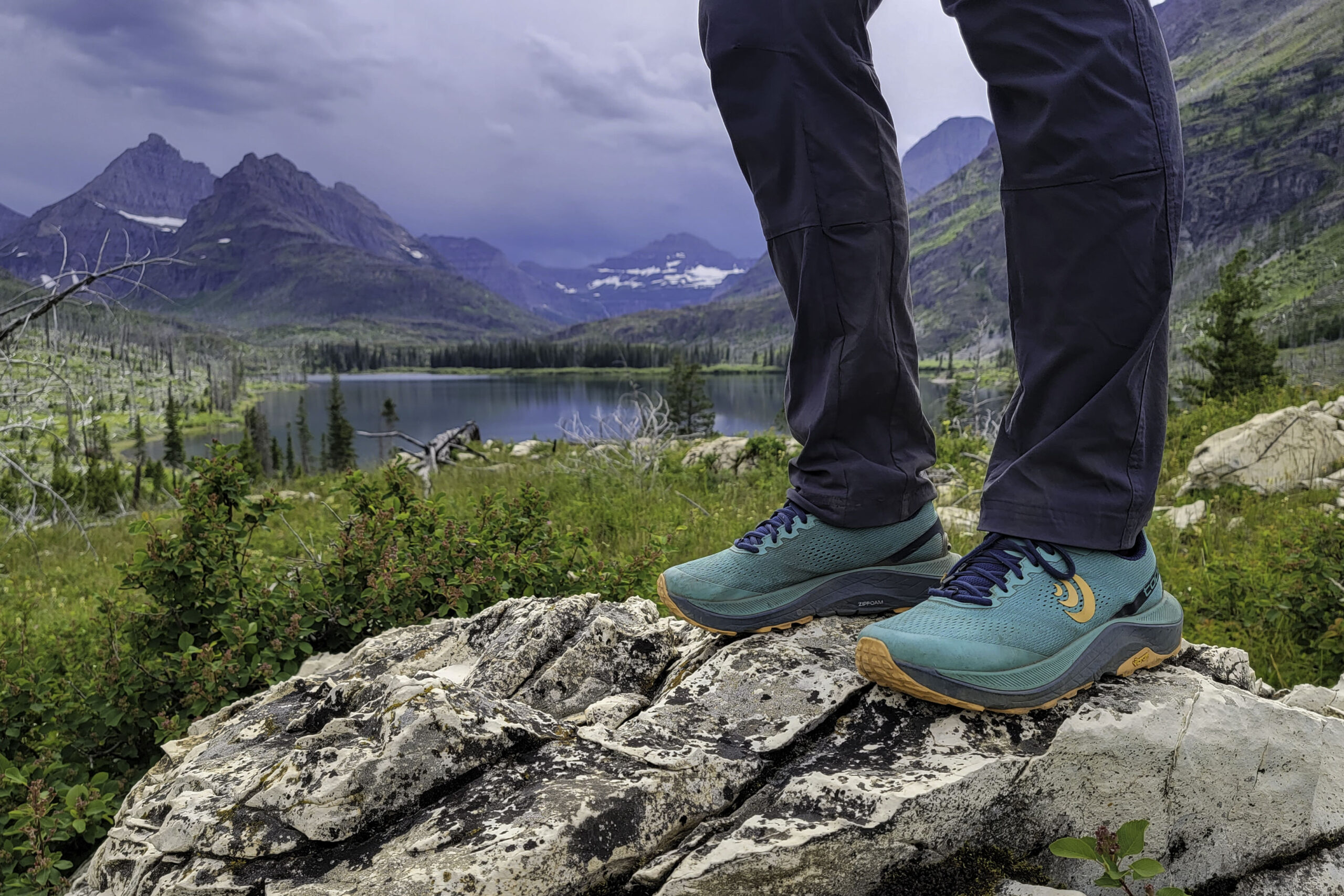 Knee-down view of a hiker standing on a white granite rock in a pair of teal Topo Athletic Ultraventure 3 hiking shoes in front a lake in Glacier National Park.