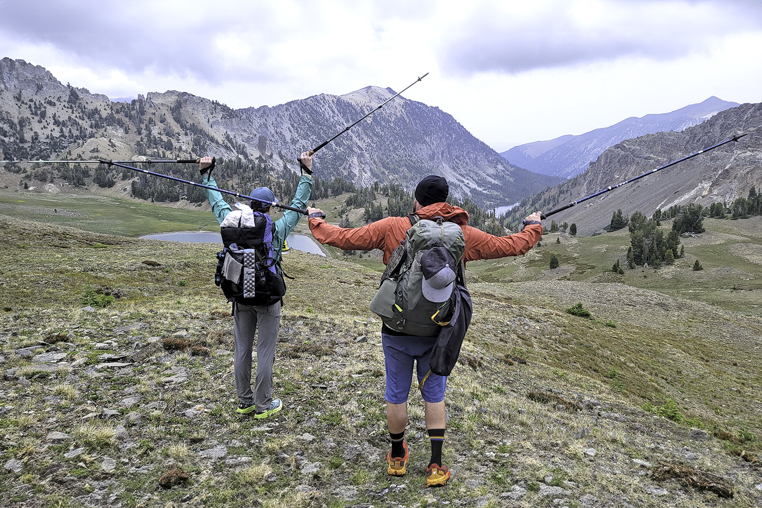 Hikers holding their Black Diamond Pursuit and LEKI Makalu Lite trekking poles up in the air while looking out over an expansive view of mountains and lakes