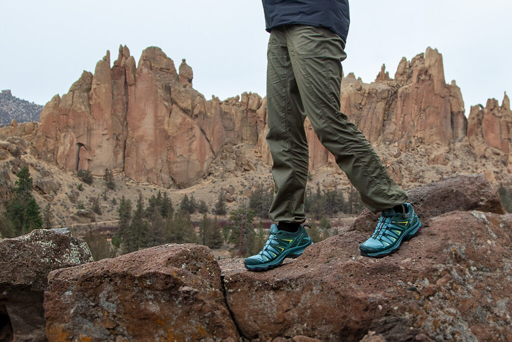 A waist-down photo of a female hiker wearing The North Face Aphrodite 2.0 hiking pants with the tuft columns of Smith Rock in the distance