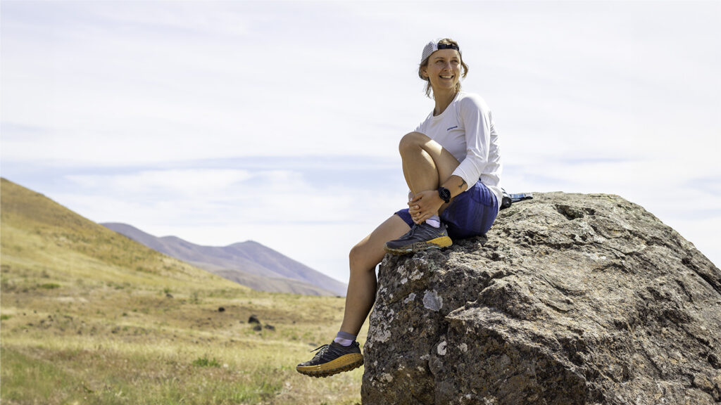 A trail runner resting on a boulder in an airy grassy hills setting in the HOKA Speedgoat 5 shoes