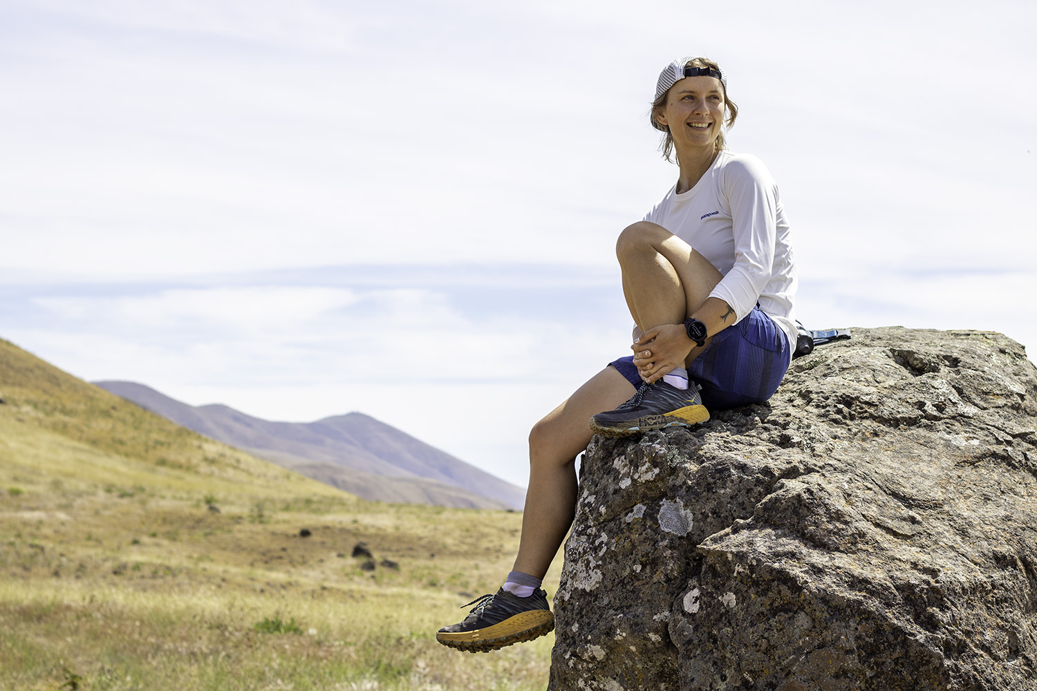 A trail runner resting on a boulder in an airy grassy hills setting in the HOKA Speedgoat 5 shoes