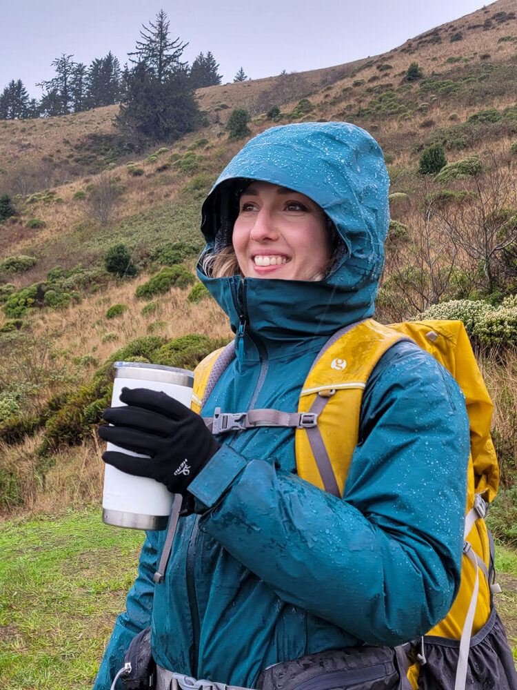 A smiling female hiker in a green Black Diamond Stormline Stretch rain jacket on a coastal hike