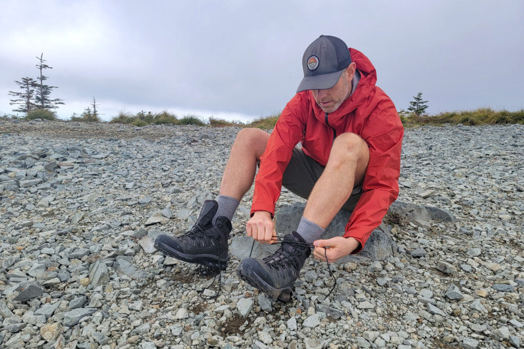 A hiker in a red rain jacket sitting on a rock to tie his Salomon Quest 4 GTX boots