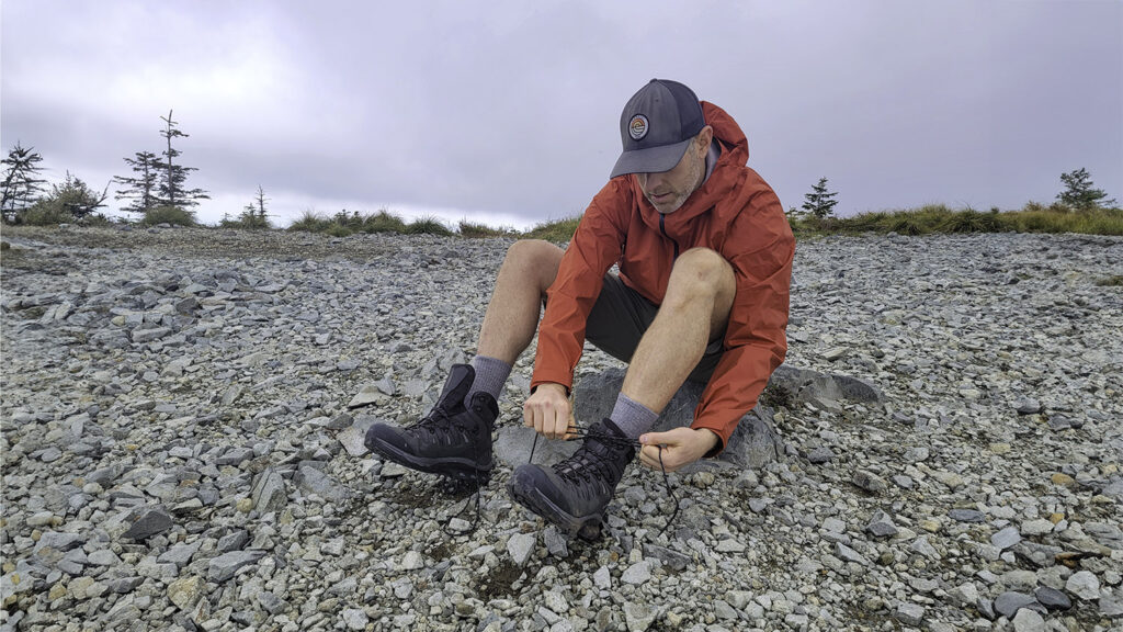 A hiker in a red rain jacket sitting on a rock to tie his Salomon Quest 4 GTX boots