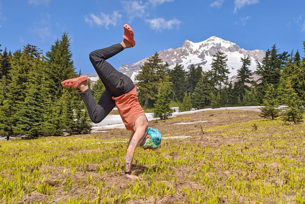 A backpacker doing a handstand in a mountain meadow in a pair of pink Crocs Classic Clogs