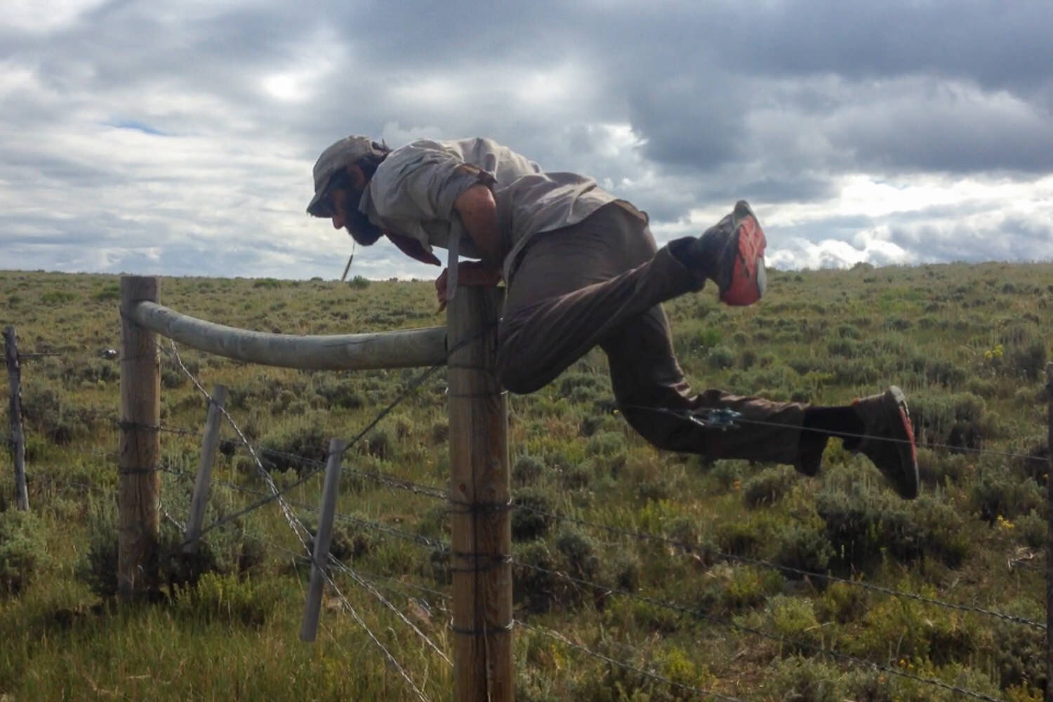 backpacker hops over a cattle fence in an open field with cloudy skies on the CDT