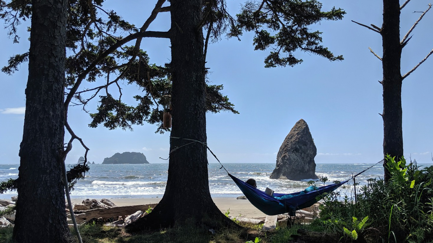 A hiker lies in a hammock looking out at a rugged coastline with crashing waves and rocks in the water.